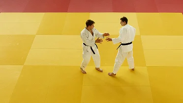 Dois atletas de judo praticando técnicas de postura e pegada em um tatame amarelo, focando em equilíbrio, posicionamento e movimento controlado durante o treinamento